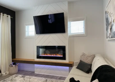 Modern living room with chevron fireplace wall, linear electric fireplace, floating wood mantel, wall-mounted TV, and light gray sectional sofa by Axium Interiors Saskatoon